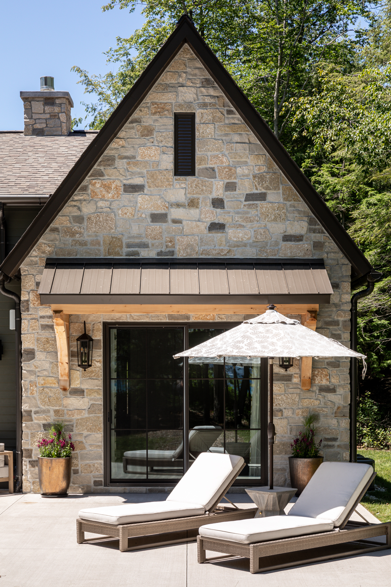 Stone cottage with a steep gable, a timber-bracketed metal awning and a patio with lounge chairs