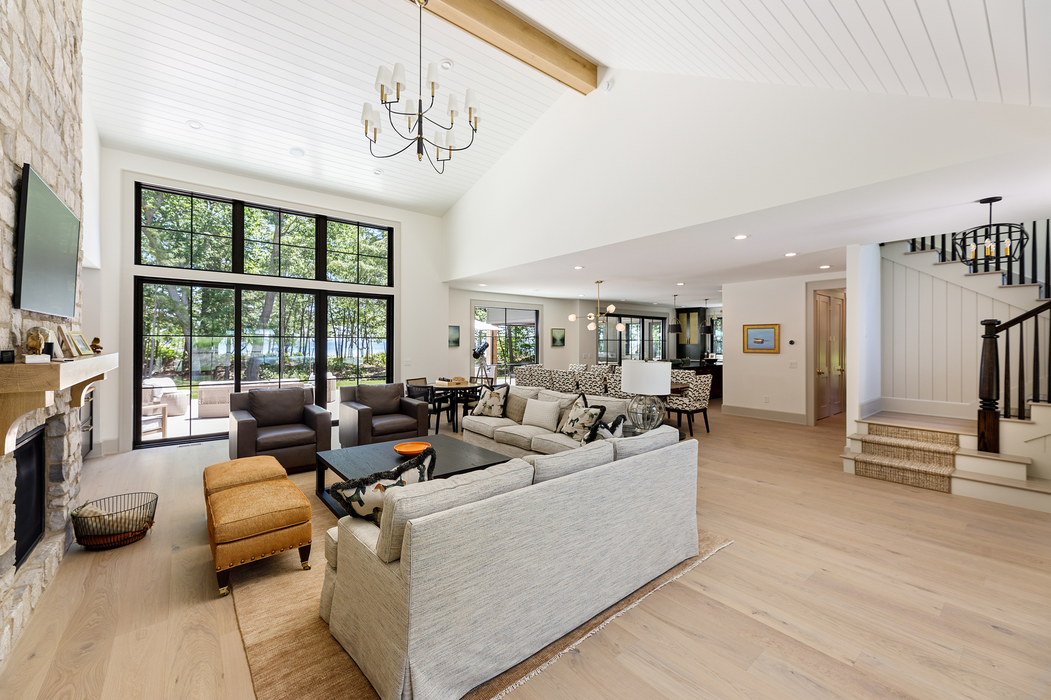 Bright great room with a white plank ceiling, black-framed windows, a stone fireplace and an open stair